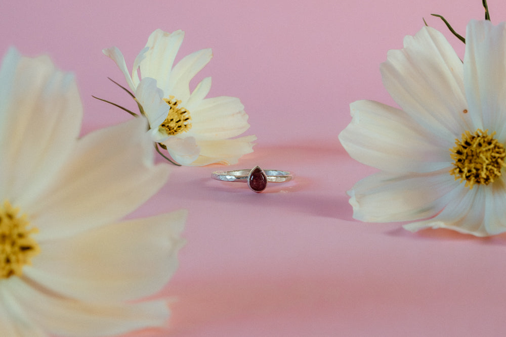 An argentium recycled silver ring with a hammered band and a teardrop shaped pink tourmaline gemstone is delicately placed on the stem of a lemon yellow cosmos flower, with another blurred flower in the background, set against a solid pink backdrop.