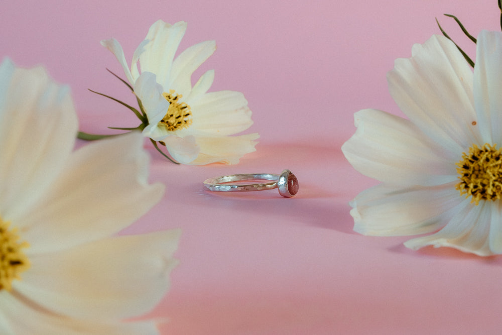 An argentium recycled silver ring with a hammered band and a teardrop shaped pink tourmaline gemstone is delicately placed on the stem of a lemon yellow cosmos flower, with another blurred flower in the background, set against a solid pink backdrop.